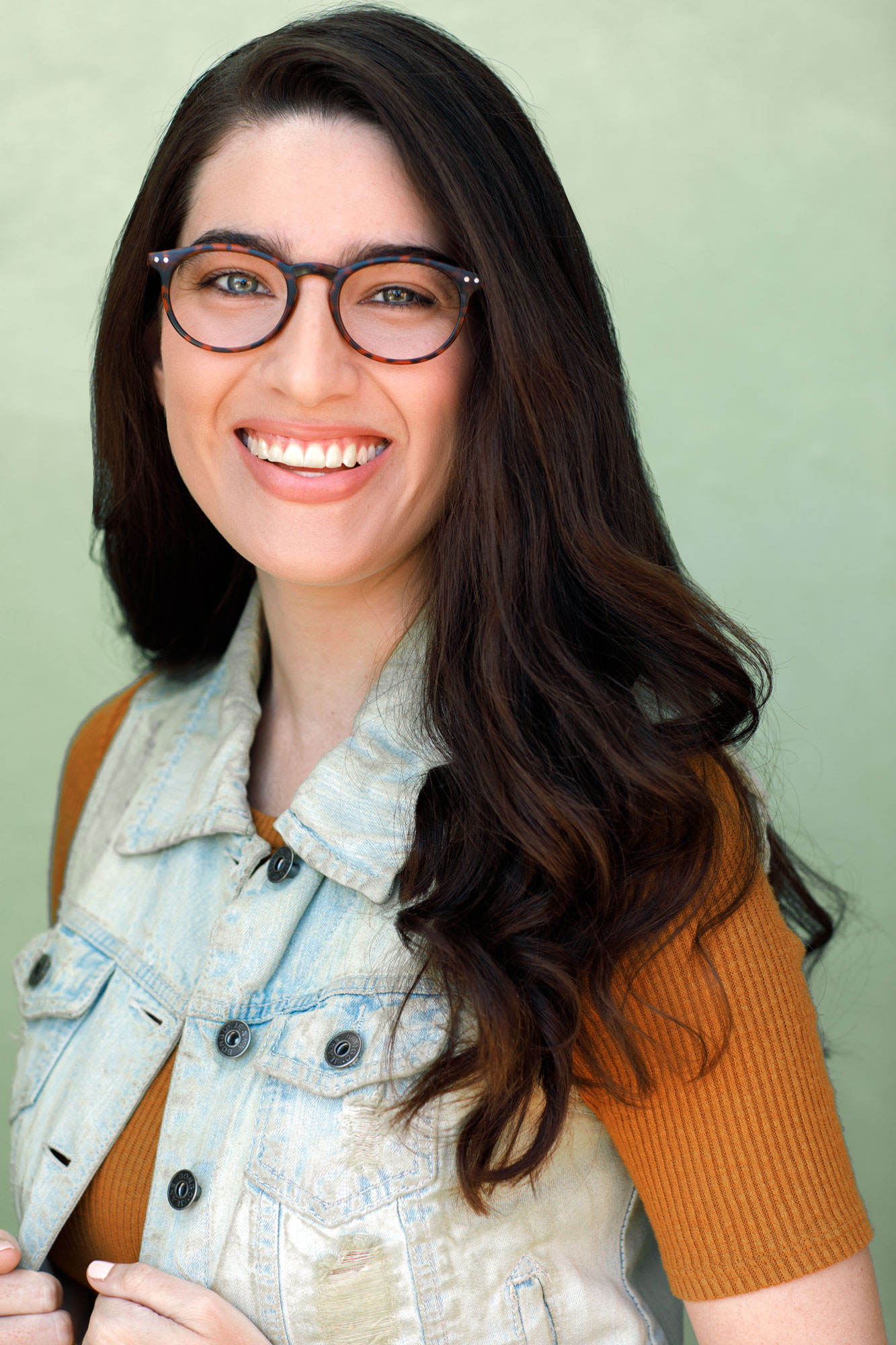 Brandy Zapata smiling with glasses wearing and orange shirt with jean jacket vest.